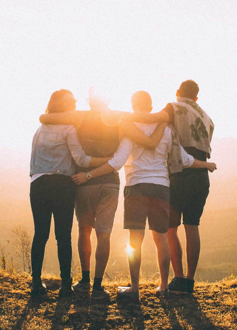 Four friends stand arm-in-arm on a hilltop at sunset, backs to the camera, gazing over a golden landscape.